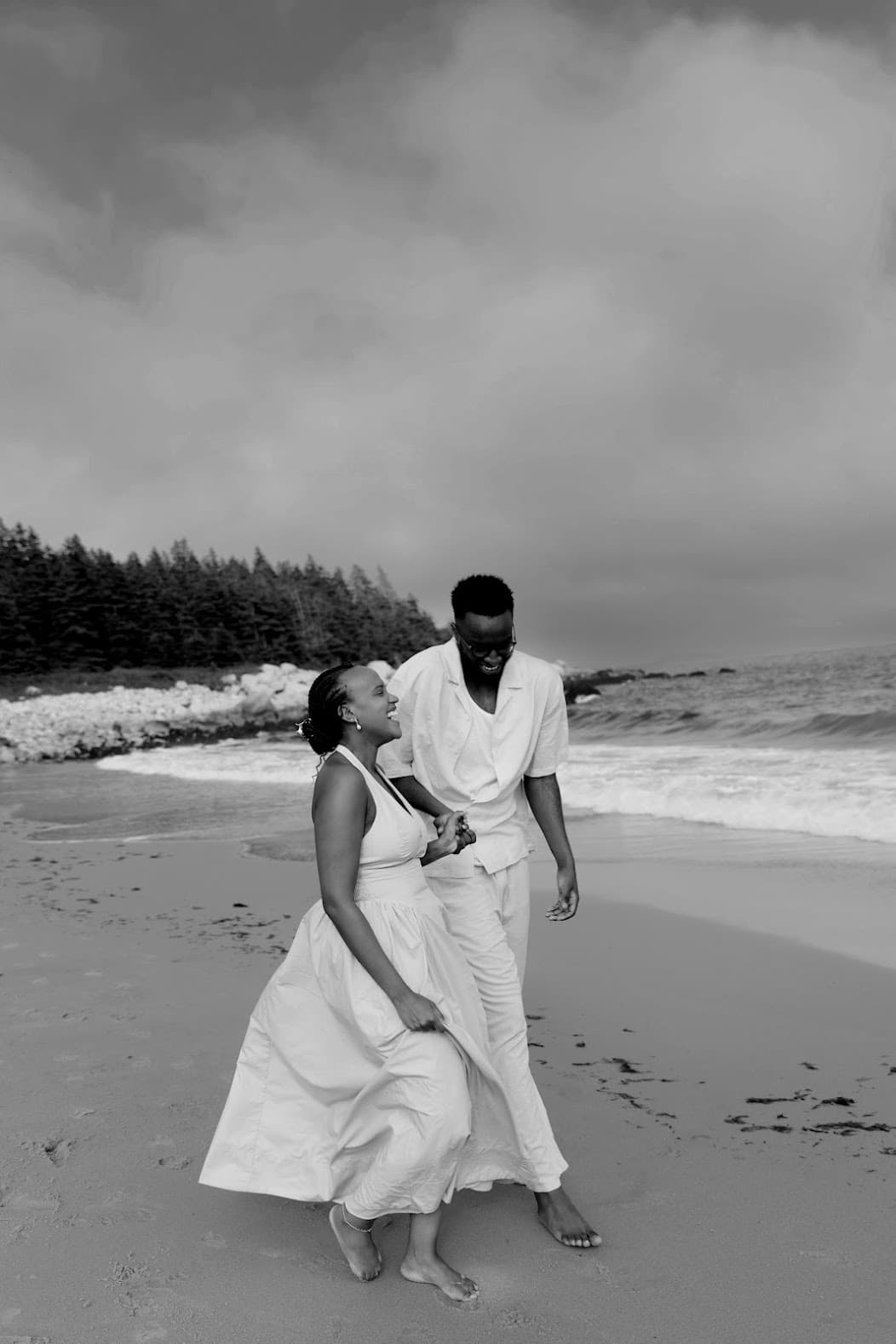 A black-and-white photograph of Joshua and Sandrine laughing together on a beach